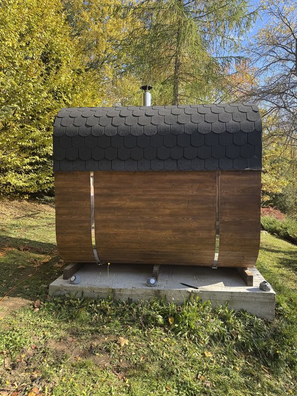 Sauna Quadro z boku, z dachem z gontu, na betonowej podstawie wśród drzew
